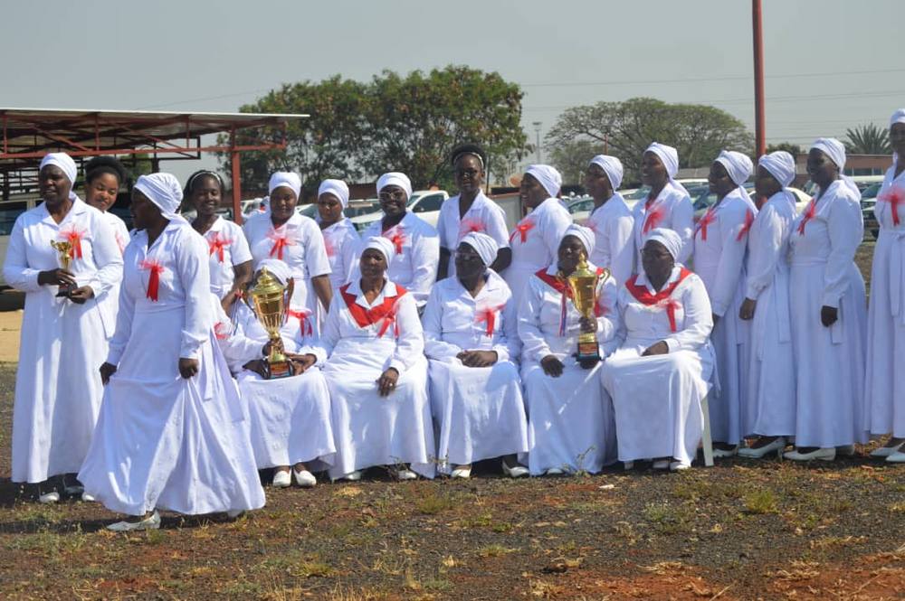 Choir members in white, seated and standing outdoors, holding three trophies